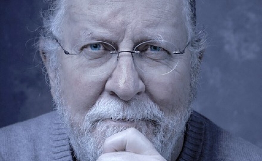 A man with grey hair and a grey beard sits with his chin propped on his hand while wearing glasses and a grey sweater.