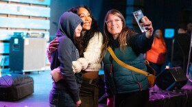 Three women pose for a selfie, one holding a phone with her arm extended and the other two embracing.