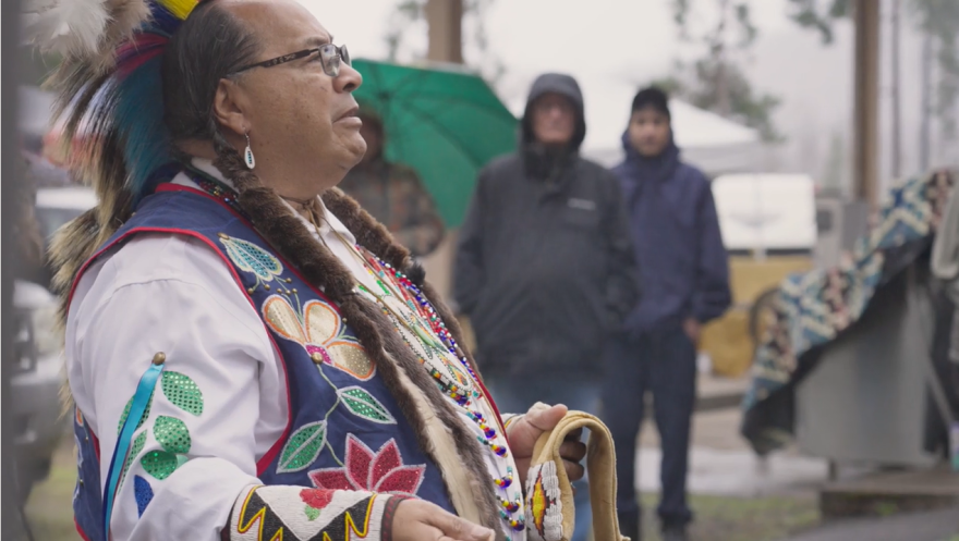 Native American man in regalia speaks to a small crowd.