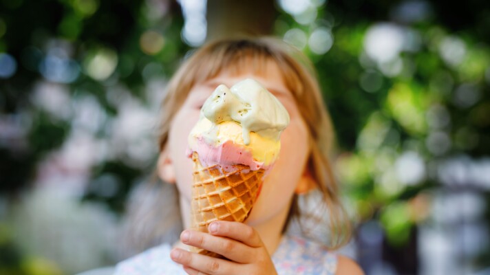 Little preschool girl eating ice cream in waffle cone on sunny summer day. Happy toddler child eat icecream dessert. Sweet food on hot warm summertime days. Bright light, colorful ice-cream.