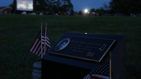 A grave marker with Harry Dean Stanton's portrait and name engraved, flanked by American flags. One of his films plays on a large screen nearby.