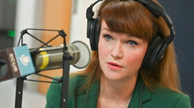 A woman sits inside a radio studio. She is sitting down and talking at a microphone.