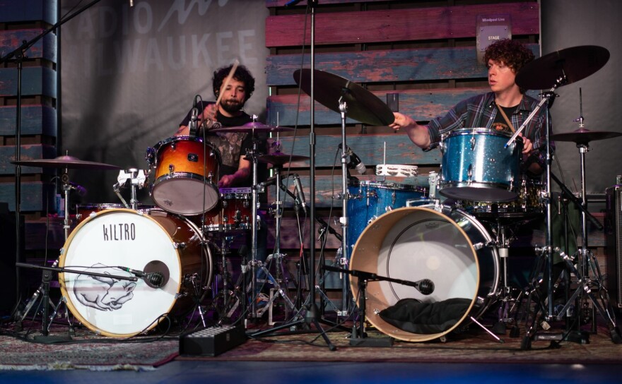  Two men play two separate drum kits on stage in a small performance space.