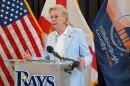 older women with short blond hair wearing a light blue pants suit with blazer. is speaking at a podium that has the Tampa Bay Rays logo. Flags of the US, Florida and Hillsborough County are behind her