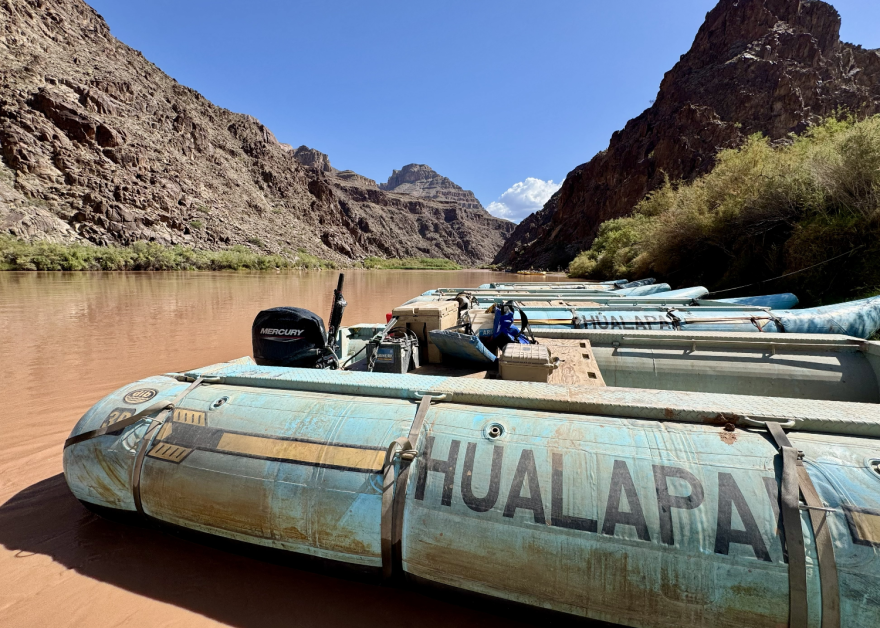 Motorized river rafts with "Hualapai" on the side sit along the banks of a brown-tinted river. There are tall rocky mountains on either side of the banks.