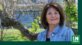 Cathy Knapp stands outdoors in front of leafy trees and a building, wearing a denim jacket, hoop earrings, and a warm expression.