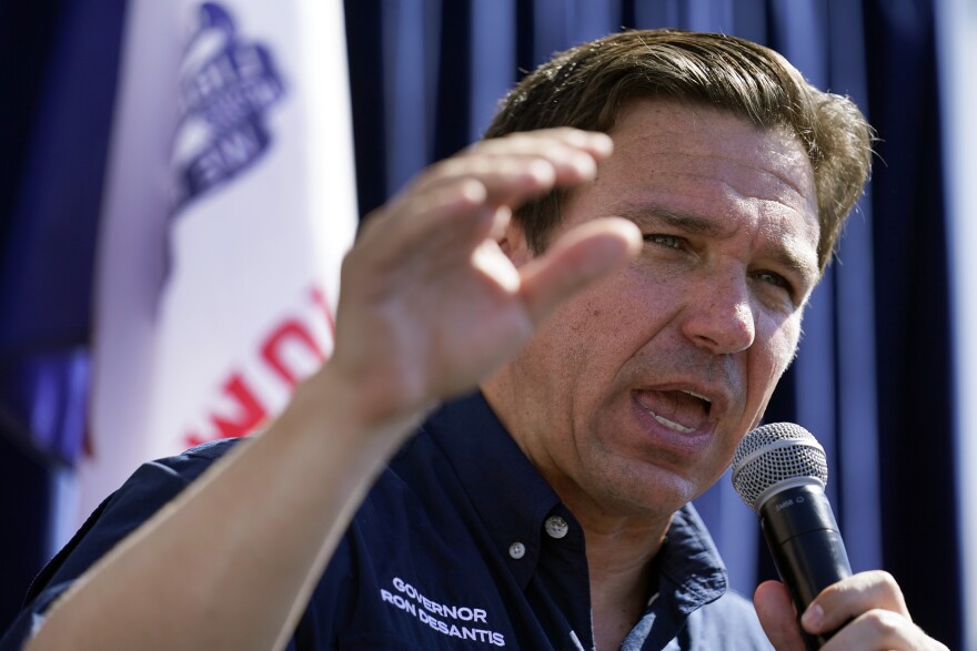A close up of a white man with short brown hair. He has a mic in his left hand and his right hand is up in the air.