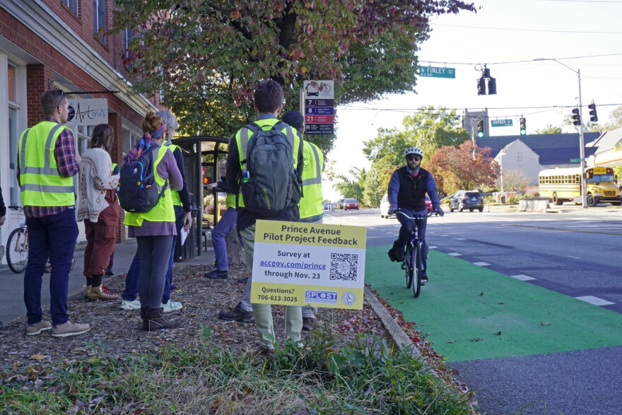 A bicyclist rides in a green bike lane along Prince Avenue while several people in safety vests stand beside a sign requesting feedback on a pilot project.