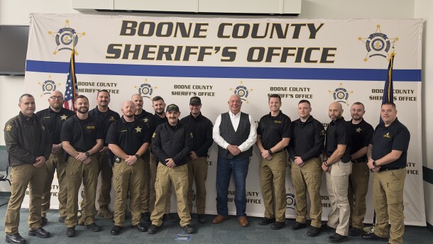 Men in black shirts and khakis pose in front of Boone County banner
