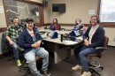 Five smiling people wearing headphones sit at a table in a radio talk studio: the man on the left in the foreground has brown hair, a brown beard and is wearing a green, black and white plaid shirt, a black t-shirt and jeans; the man on the right in the foreground has a brown beard and is wearing a black baseball cap, a blue sweatshirt, a white t-shirt and jeans; the woman on the left in the background has shoulder length dark hair and is wearing glasses, a red and black plaid long sleeved shirt and a red t-shirt; the woman on the right in the background is wearing a purple hat, glasses and a blue t-shirt; the man on the end has short brown hair and a brown beard and is wearing glasses, a white button down shirt, a blue sweatshirt and jeans.