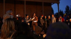The Vice Mayor of Truckee dressed in a brown coat speaks into a microphone to a group of people outdoors in the evening at a vigil.