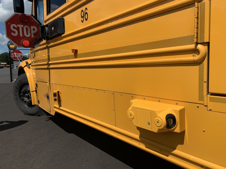 Left side of a school bus with its red and white stop arm opened. Midway along the bus and 36" off the ground is a metal box with a camera tucked inside.