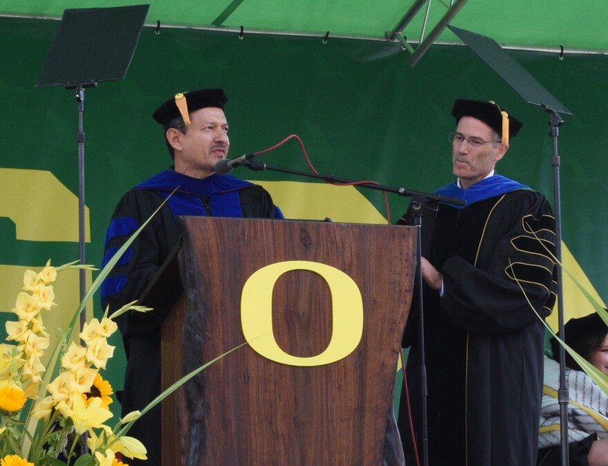 A person speaking at a podium. The person is dressed in academic regalia. The podium is adorned with a large O. A person is standing next to the podium, also dressed in academic regalia.