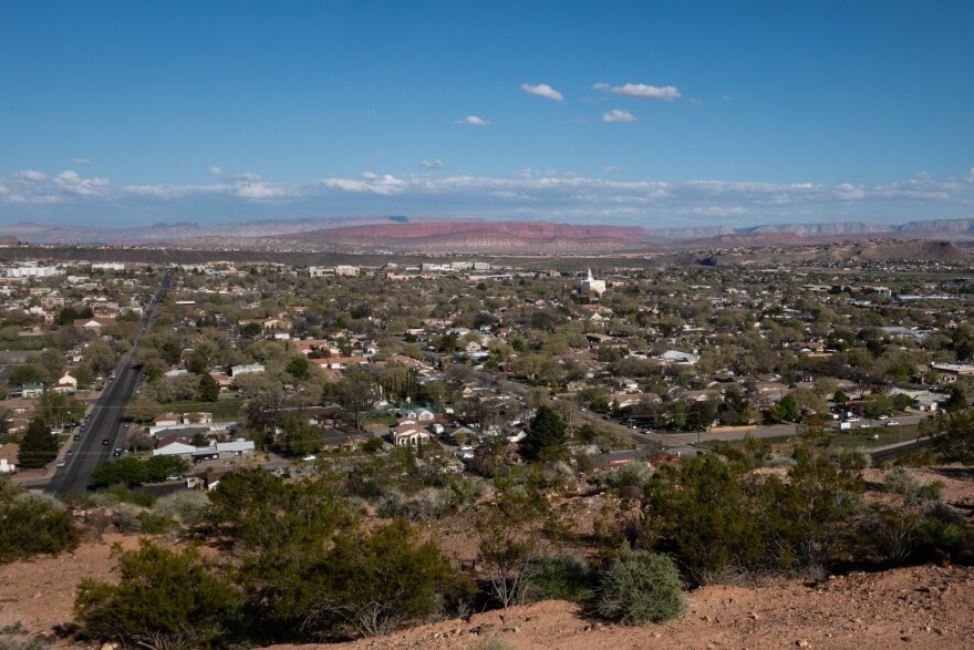  The city of St. George, seen from above. A grid of rooftops, treetops stretch out towards red cliffs in the distance. 