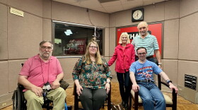 Five people pose for a photo in a sound-proof tan radio studio. On the far right, sitting in a blue frame manual wheelchair is Todd Vaarwerk, a white man with grey beard and short hair. He's wearing glasses, a pink polo shirt and tan pants. To Todd's right, also sitting is Stephanie Orlando, a white woman with long blonde hair with side bangs. She is wearing a green print blouse, dark pants and purple glasses. Next to Stephanie, also sitting, is Craig Donatelli, a white man with Down syndrome. He has short brown hair, is wearing round glasses, a Captain America shirt, and jeans. Behind Craig, standing, are his parents Joyce and Max. Joyce is on the left and has shoulder length blonde hair and is wearing a pink top, pink fleece jacket with black pants. Max stands to the right of her with his arm around her. Max has short white-grey hair and a beard. He's wearing a mint green and black stripped polo shirt and jeans. 