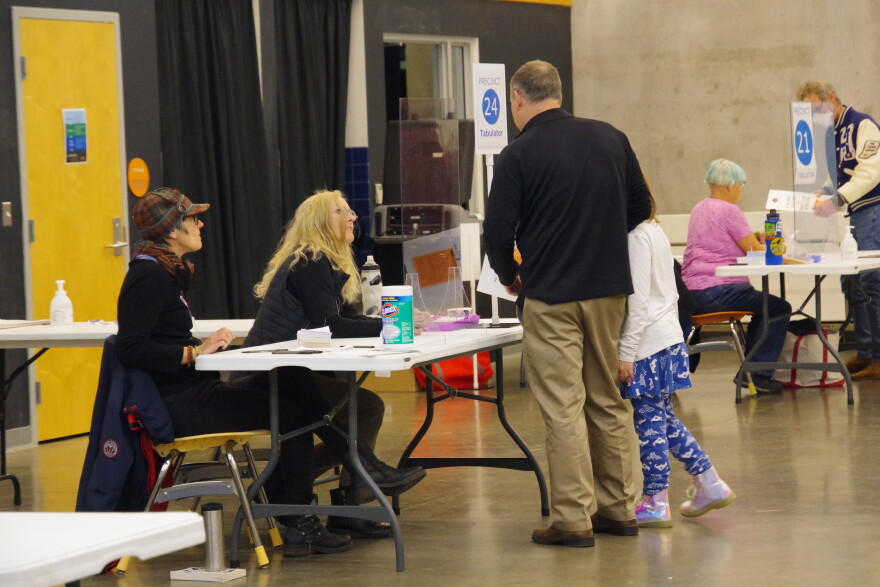 A man in a black shirt and tan pants takes his daughter to a precinct in the Girl Scout headquarters in Kalamazoo.  He's holding his ballot, talking to poll workers as he gets ready to put his completed ballot into the voting machine. 