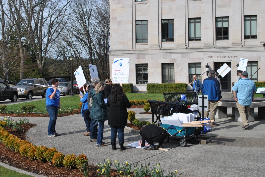 People wearing coats and jeans are gathered in front of a stone building holding picket signs. Someone is kneeling over a poster on the ground as they draw out a message.