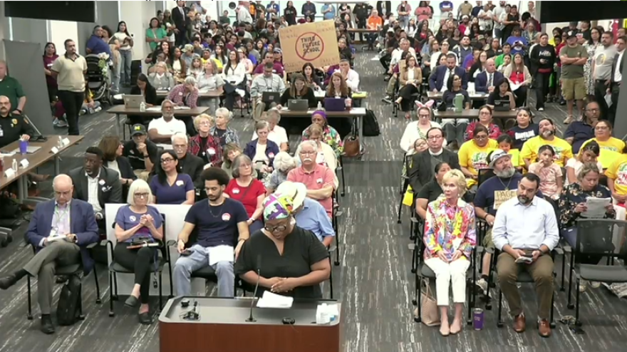 A woman in a headscarf and glasses speaks at a podium with a full boardroom behind her, including someone holding a sign that says "Third Future Schools" with a line through it.