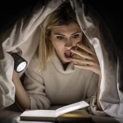 Beautiful young woman reading book under bed cover and holding a flashlight.