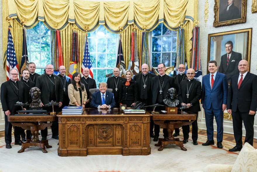 Bishops and White House officials stand around President Trump in the oval office and pose for a photo. Flags and gold curtains hang behind them.