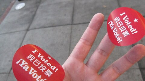 A photo of a hand with light skin holding two stickers from San Francisco that are red with white text. They say "I voted" in English, Chinese and Spanish. 