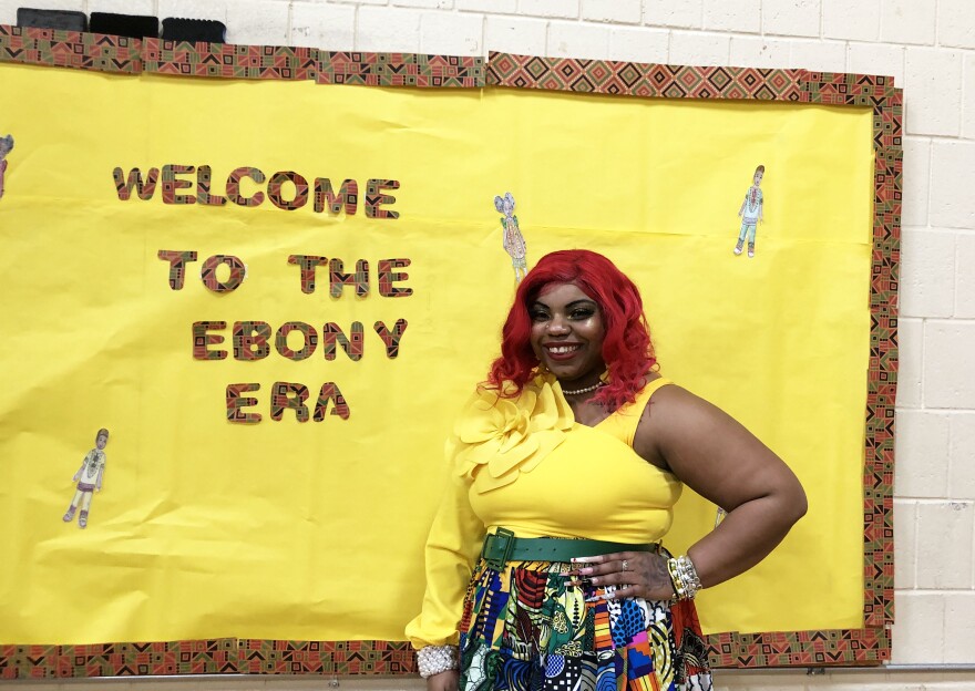 A woman stands in front of a bulletin board with the message "Welcome to the Ebony Era" displayed on a yellow background. 