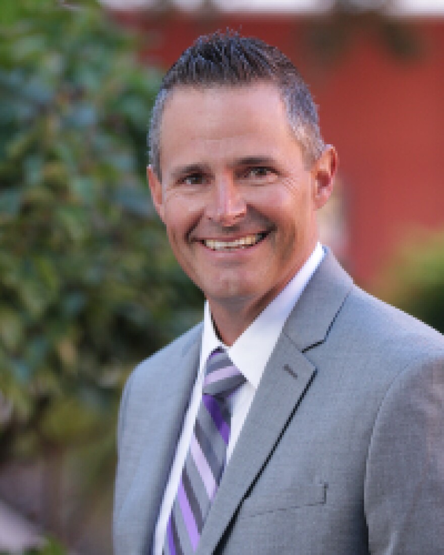 Man in a suit stands in front of a background with blurred shrubbery.