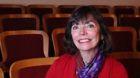 A white woman with dark hair smiling at the camera while seated in a theater. She is wearing a red sweater set and a multi-colored silky scarf around her neck.