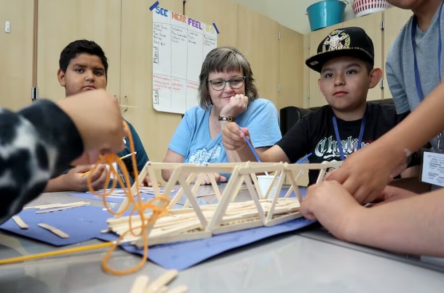 Students' hands work on a bridge made out of popsicle sticks. In the background, two other students watch and contribute to the structure. In between them, a woman watches with her head perched on one hand.