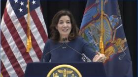 Gov. Kathy Hochul, wearing a blue dress speaking at a podium with U.S. and state flags behind her.