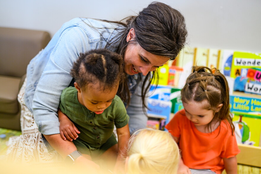 A woman smiles as she interacts with three young children in a classroom, surrounded by colorful books and educational materials.