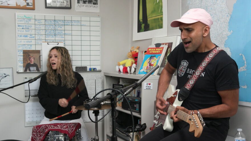 Shahjehan Khan plays guitar while wearing a Brown University T-shirt and a pink baseball cap. Talia Temporal plays drums in the background while wearing a black sweater. Both musicians are singing back-up vocals while playing their instruments.