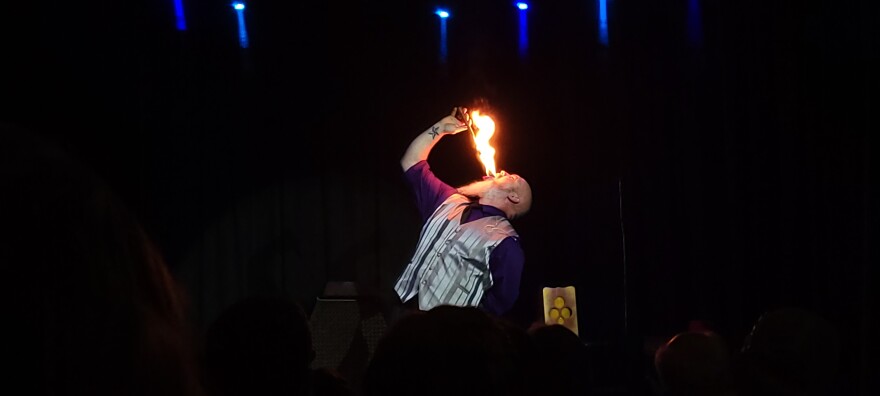 A man onstage puts a flaming baton into his mouth.