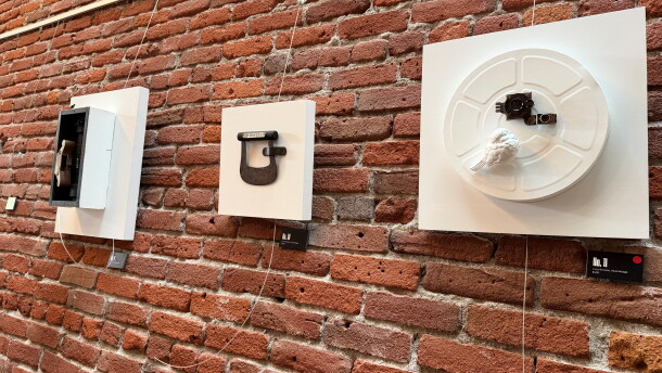 A brick wall with three white boards hanging from it. Each has found objects connected to them as examples of assemblage art.