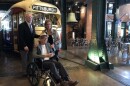 A man and woman stand with a man in a wheelchair in front of a vintage trolley car inside a museum.