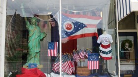 A storefront has a mini Statue of Liberty and many American flags in the window, including several flags that say "250" or '76. The largest flag is an Ohio flag with "250" in the center of the circle. A mannequin wears an America 250 shirt and a skirt made out of red, white, and blue bunting. 