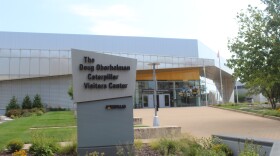 Exterior of the Doug Oberhelman Caterpillar Visitors Center, a modern building with angular, sail-like metal paneling and a large monument sign reading "The Doug Oberhelman Caterpillar Visitors Center" alongside the Caterpillar logo. Glass doors and windows with photo displays are visible under a bright yellow awning band.