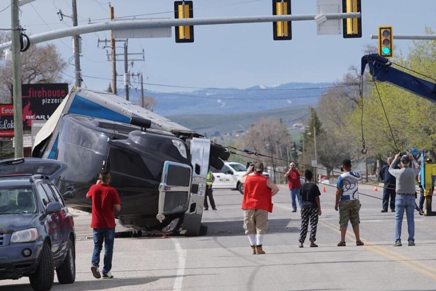 A semi-truck lies on its side in the middle of a road. People stand nearby watching as cords are attached to the wheels.