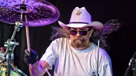 Man with beard and long hair wearing glasses and white tshirt drumming on stage
