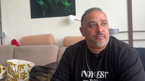 Owen Ramsingh sits in his father's house in the Netherlands. He's wearing a black tee shirt and sitting on a couch with white pillows that have a yellow leaf stencil on them.