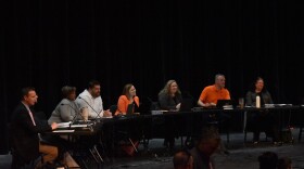 Seven people seated at table with microphones in front of them and a black backdrop behind them.