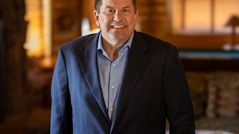 A man wearing a dark blazer smiles for the camera. He’s standing inside a log cabin room.