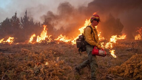 A firefighter holds a fuel canister as a prescribed fire burns in the background.