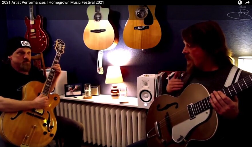 Video still of a man playing guitar with another man who is also him