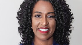 A smiling woman with curly black hair with her arms crossed in front of her chest. She is wearing a gold necklace, a pink top, and a blue blazer. 