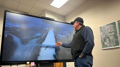 Lincoln County Sheriff Darren Short points at a large screen showing an aerial image of a bridge in Libby, MT. Short spoke about the flood damage at a press conference in Libby on December 12, 2025.