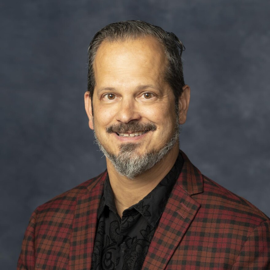 A smiling man with a dark red checkered jacket and a black shirt looks toward the camera.