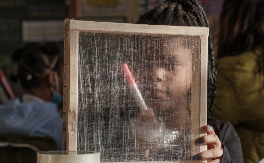 A Black child seen through a screen in a square wooden frame.