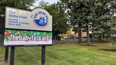 Sign that reads 'The Baby Fold, serving children and families since 1902' and 'thebabyfold.org' outside a building lined with trees 