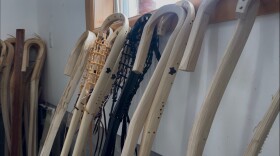 Rows of shorn hickory sticks with leather netting bound around a bent end stand against a windowsill waiting for further processing as future hand-hewned lacrosse sticks.